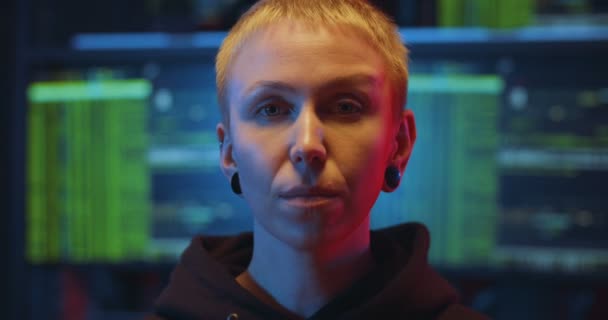 Portrait of Caucasian woman hacker with short hair looking straight to camera and chewing gum in monitoring room at night. Close up of female cyber criminal with big screen on background. — Stock Video