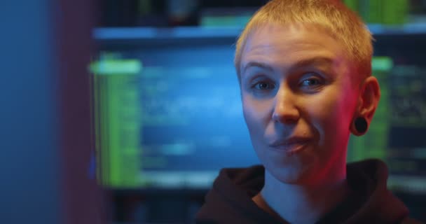 Portrait de hacker femme audacieuse caucasienne aux cheveux blonds courts regardant droit devant la caméra et mâchant de la gomme dans la salle de surveillance la nuit. Gros plan cybercriminel féminin audacieux avec écran en arrière-plan . — Video