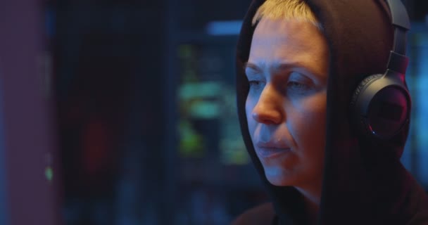 Close up of Caucasian woman in hood and headphones committing cybercrime and looking attentively at computer screen at night. Female hacker hacking system and program online in dark room. — Stock Video