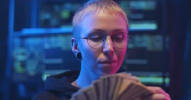 Gözlüklü beyaz kadın hacker 'ın karanlık odada bir sürü monitörle para saydığını gördüm. Kadın siber suçlu, elinde dolarlarla elinde banknotlar tutuyor. Kirli maaş ve çamaşırcılık konsepti.