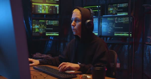 Běloška v kápi a sluchátka páchající kyberkriminalitu, pozorně se dívající v noci na obrazovku počítače a pak se usmívající. Žena hacker krást data ze systému v tmavé místnosti s monitory. — Stock video