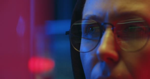 Gros plan des yeux dans les lunettes de femme caucasienne grave à la cagoule travaillant et regardant attentivement l'écran d'ordinateur la nuit dans le bureau informatique. Fraude de pirates sur Internet. Cyber policier . — Video