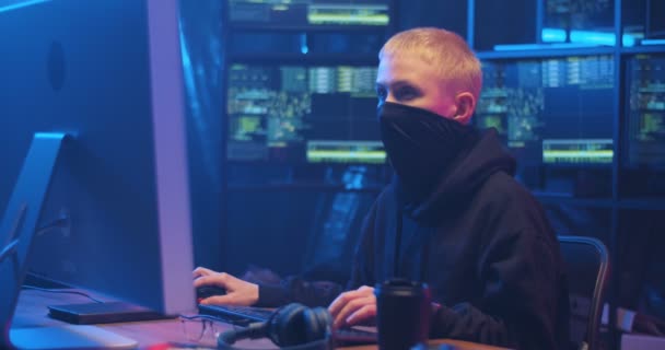 Caucasian woman hacker with blond short hair and masked face sitting in dark room with monitors and typing on keyboard of computer. Female hacking online system. Stealing information concept. — Stock Video