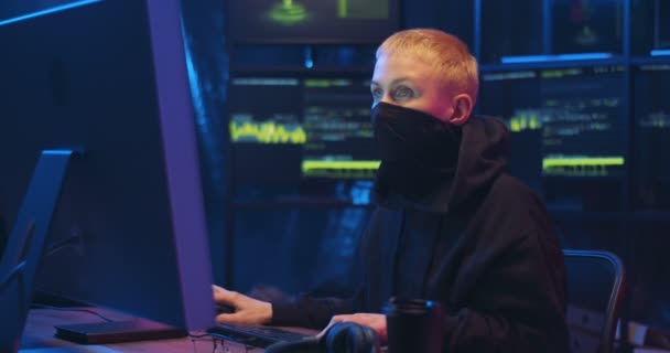 Hacker mujer caucásica con el pelo corto y rubio y la cara enmascarada sentado en la oscuridad delante de la computadora. Mujer enmascarada trabajando en ciberseguridad. Concepto de ladrón de datos digitales . — Vídeo de stock