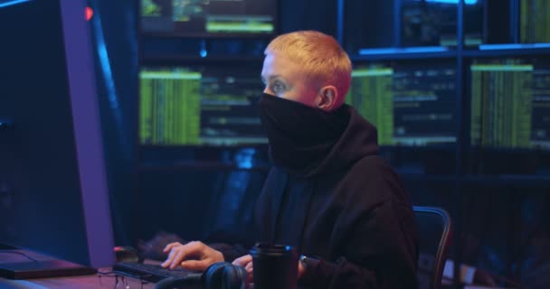 Caucasian woman cyber thief with blond short hair and mask on face sitting in darkness at desk and typing on keyboard of computer. Anonymous female hacking system. Laundring money online concept. — Stock Video