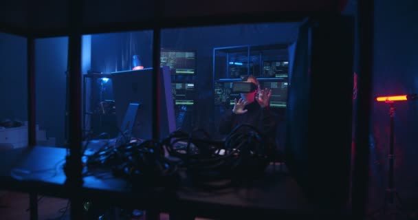 Biała kobieta haker w okularach Vr siedzi w ciemnym pokoju pełnym komputerów i ma wirtualne słuchawki. Kobieta to pracownik stukanie i przewijanie w powietrzu. — Wideo stockowe