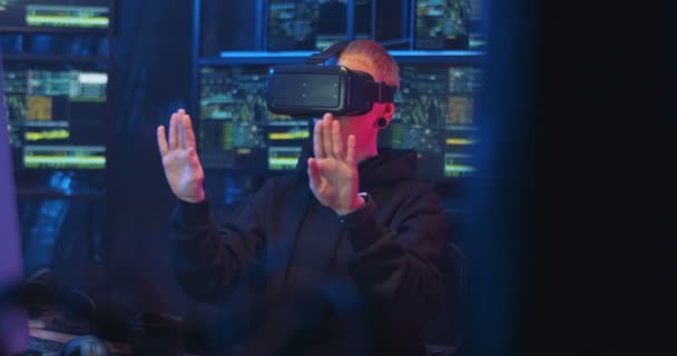 Hackeadora caucásica en gafas VR sentada en la oscuridad en la sala de monitoreo llena de computadoras mientras tiene auriculares virtuales. Mujer trabajadora de TI tocando y desplazándose en el aire. Mujer policía cibernética . — Vídeos de Stock