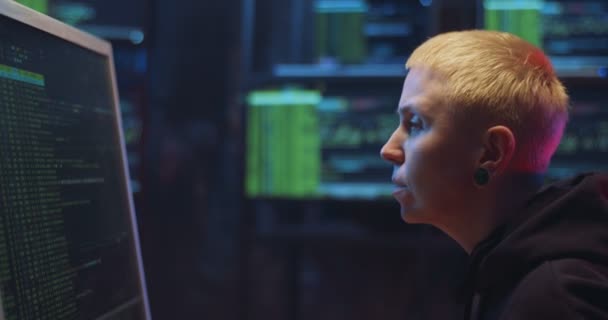 Side view on Caucasian woman hacker with blond short hair sitting in dark room with monitors at desk and working on computer with digits. Female hacking online system. Stealing information concept. — Stock Video