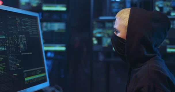 Visão lateral da mulher caucasiana com rosto mascarado e no capuz trabalhando no centro de segurança cibernética e analisando dados. Feminino hacker anônimo digitando no teclado no computador na sala de monitoramento escuro . — Vídeo de Stock