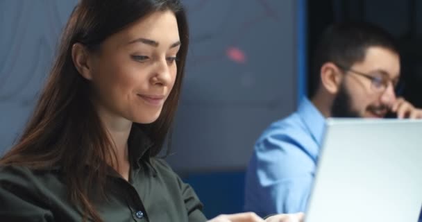 Mulher morena muito caucasiana sorrindo ao trabalhar no computador portátil no escritório no turno da noite. Macho colega de trabalho em óculos falando no telefone em fundo embaçado. Trabalhadora bonita no trabalho . — Vídeo de Stock