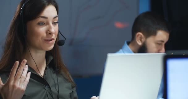 Vit vacker brunett ung kvinna i hörlurar och mick har videochatt på bärbar dator i callcenter. Kvinnlig arbetare på callcenter pratar med klienten på kvällen. Manlig medarbetare bakom. — Stockvideo