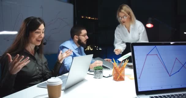 Bella giovane donna caucasica che lavora al computer nel call center e parla con il cliente tramite web cam. Lavoratrice di call center a tarda sera. Colleghi che parlano e discutono dietro . — Video Stock