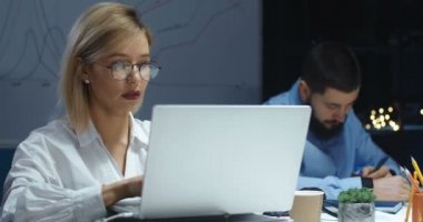 Akşamları bilgisayar başında çalışan güzel, beyaz, sarışın bir kadın. Dizüstü bilgisayarda yazan ve mesaj atan bir kadın. Arka planda oturan bir erkek arkadaş. Masadaki işçiler geç saatlere kadar çalışıyor..
