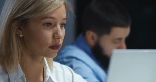밤늦게 사무실에 있는 랩탑 컴퓨터에서 일하는 아름다운 금발의 젊은 여성 이 가까이 있습니다. 예쁜 여성 회사 직원 이 화면을 타이핑하고 보고 있습니다. 배경 이 흐릿 한 남성 동료. — 비디오
