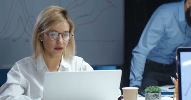 Mujer rubia joven que trabaja hasta tarde en la noche en el ordenador portátil. Mujer bonita escribiendo y mensajes de texto en el ordenador portátil. Hombre compañero de trabajo que viene y se sienta en la mesa en el fondo . — Vídeo de stock