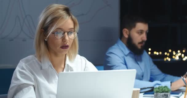 Mujer rubia joven caucásica trabajando y escribiendo en el ordenador portátil. Una trabajadora guapa en la oficina tarde en la noche. Hombre compañero de trabajo haciendo su trabajo sobre fondo borroso . — Vídeos de Stock