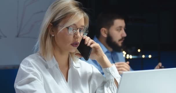 Ung vit vacker och leende blond kvinna i glasögon talar på mobiltelefon medan du skriver på bärbar dator. Kvinnlig kontorsanställd som talar i mobiltelefon på kvällen. Manlig kollega bakom. — Stockvideo