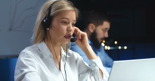 Vit vacker blond ung kvinna i hörlurar med mick har videochatt på bärbar dator i callcenter. Kvinnlig arbetare på callcenter pratar med klienten på kvällen. Manlig medarbetare bakom. — Stockvideo