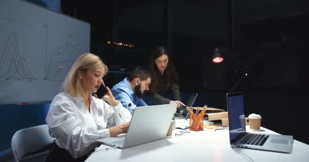 백인젊은 금발 여성 이 휴대폰으로 통화하고 야간 교대로 노트북 컴퓨터에서 일하고 있습니다. 남성 과 여성의 회사 동료들 이 배경에 협력하고 있습니다. 휴대폰으로 통화하는 여자. — 비디오