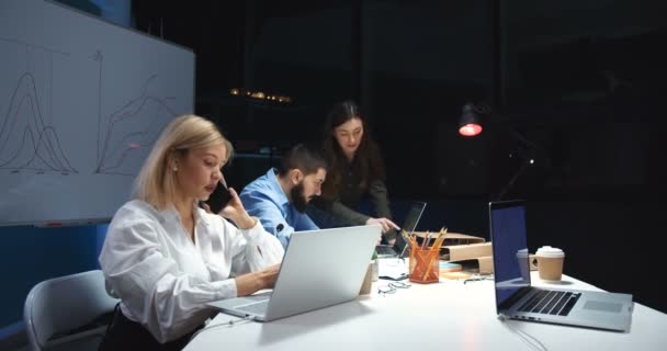 Mujer rubia joven caucásica hablando por teléfono móvil y trabajando en el ordenador portátil en la oficina en el turno de noche. Colaboradores de empresas masculinas y femeninas que cooperan en sus antecedentes. Señora hablando por celular . — Vídeos de Stock