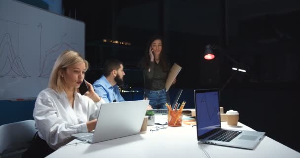 Biała młoda blondynka rozmawiająca przez telefon komórkowy i pracująca na laptopie w biurze na nocnej zmianie. Współpracownicy firmy męskiej i żeńskiej współpracujący w terenie. Pani rozmawia przez komórkę.. — Wideo stockowe