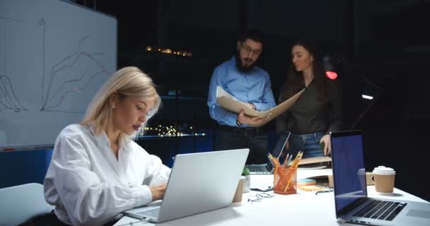 会社の男性と女性の共同作業者は、夕方遅くまで忙しいとしてオフィスで一緒に働いています。ブロンドの女性はテーブルに座って、ラップトップコンピュータを使用しています。男性と女性の同僚は文書を読んで話す. — ストック動画
