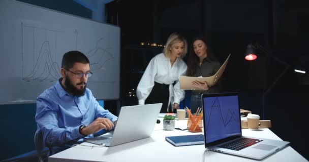 Hombre caucásico en gafas de trabajo en el ordenador portátil por la noche. Dos hermosas compañeras de trabajo hablando y discutiendo algún proyecto mientras leen documentos y escriben a bordo . — Vídeos de Stock