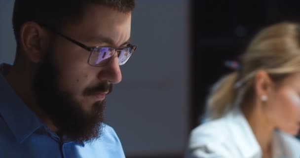 Närbild av kaukasisk man i glasögon med allvarligt ansikte och svart skägg tittar på datorskärmen när du arbetar på kontoret på kvällen. Man upptagen arbetare i företaget. Blond kvinna på suddig bakgrund. — Stockvideo
