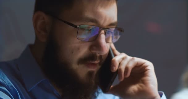 Close up de homem caucasiano de óculos com rosto sério e barba preta falando no telefone celular enquanto trabalhava no escritório à noite. Trabalhador da empresa a falar ao telemóvel. Conceito de chamada de negócios . — Vídeo de Stock
