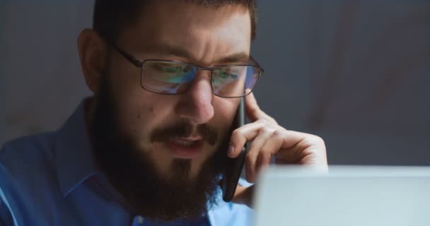 Detailní záběr bělocha v brýlích s vážným obličejem a černým vousem, jak mluví na mobilním telefonu před obrazovkou notebooku. Mužský pracovník společnosti, který mluví na mobilu. Koncept obchodního hovoru. — Stock video