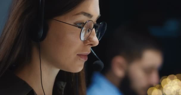 Close up of beautiful young Caucasian brunette woman in glasses, headphones with mic working late evening in office and looking at screen. Female worker of call center. Male colleague on background. — Stock Video