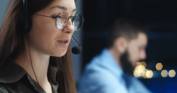 Närbild av vackra unga kaukasiska kvinna i glasögon, hörlurar med mikrofon pratar med klienten på natten och tittar på skärmen. Kvinnlig arbetare på callcenter ler mot kameran. Manlig medarbetare bakom. — Stockvideo