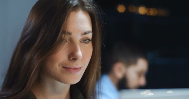 Närbild av vackra unga kaukasiska kvinna som arbetar sent på kontoret på datorskärmen. Kvinnlig företagsarbetare ler glatt och blinkar ett öga. Manlig medarbetare bakom. Porträtt. — Stockvideo
