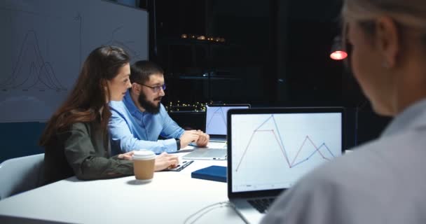 Colaboradores caucásicos, hombres y mujeres, trabajando juntos en un proyecto empresarial. Mujer bebiendo café y hombre tocando en el teléfono inteligente en la oficina tarde en la noche. Colegas hablando de gráficos y gráficos . — Vídeo de stock