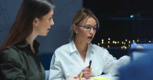 Unga attraktiva kvinnor arbetar tillsammans på kontoret sent på kvällen medan de diskuterar affärsprojekt på bärbar dator. Kvinnliga kollegor brainstormar tillsammans på företagsmöte. — Stockvideo