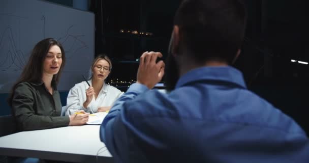 Parte trasera del jefe masculino caucásico sentado en el ordenador portátil y en la mesa con dos hermosas trabajadoras de la compañía mientras describe y cuenta la estrategia de negocios. Vista posterior del hombre hablando con las mujeres en la oficina . — Vídeos de Stock