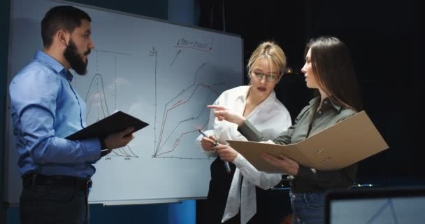 Donna bionda caucasica che disegna a bordo alcuni numeri grafici e di scrittura mentre lavora con il team in carica a tarda sera. Uomo e due donne che creano business plan per la start-up . — Video Stock