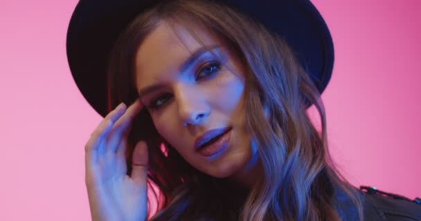 Veselý stylový brunetka model v klobouku při pohledu do kamery ve studiu v neonově modrém světle. Ženská módní střelba. Detail portrét ženy na růžovém pozadí. — Stock video