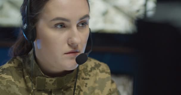 Porträtt av vit ung kvinna i headset sitter på datorskärmen i övervakningsrummet i armén och tittar sedan på kameran. Närbild av kvinnliga soldat sändare i militärt centrum. — Stockvideo