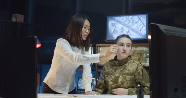 軍の管理事務所で勉強する兵士の制服を着た若い白人の少女。アジアの女性教師は軍隊で女性を教え、監視室でコンピュータ画面に何かを示す. — ストック動画