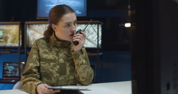 Caucasian young woman in camouflage uniform sitting at desk in front of computer screen in monitoring room and talking in walkie-talkie. Female soldier using radio transmitter, command center. — Stock Video