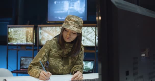 Mujer joven asiática soldado en uniforme y gorra sentada en el escritorio frente a la pantalla de la computadora y escribiendo algunos documentos en la sala de monitoreo del ejército. Mujer trabajando en centro de control militar . — Vídeo de stock