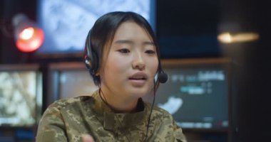 Üniformalı ve kulaklıklı Asyalı genç bir kadın ordudaki kontrol odasında bilgisayar ekranında oturup video şapkayla konuşuyor. Askeri veri merkezinde çalışan kadın askere yakın çekim.
