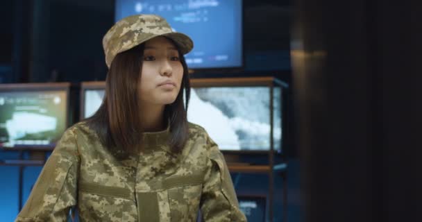 Portrait of beautiful Caucasian young woman in camouflage uniform and hat sitting at desk and working at computer, then turn face to camera and looking straight Female in military monitoring center — Stok Video