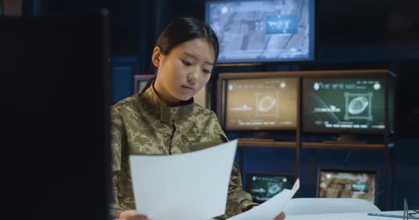 Ázsiai fiatal csinos nő egyenruhában ül az asztalnál a számítógép képernyője előtt, és iratokat ír a hadsereg megfigyelő termében. Katonai központban dolgozó nő. — Stock videók