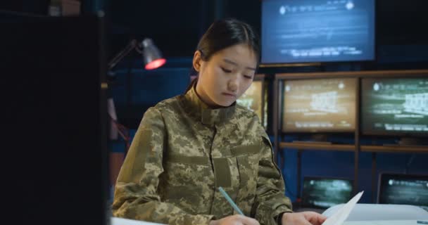 Yong asiática hermosa mujer en camuflaje sentado en la mesa delante de la pantalla de la computadora y escribir algunos documentos en la sala de vigilancia de las tropas. Mujer trabajando en centro de control militar . — Vídeo de stock