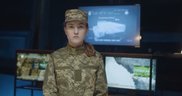 Portret van een serieuze blanke vrouwelijke soldaat in uniform en pet die in de controlekamer staat en recht naar de camera kijkt met schermen op de achtergrond. Jonge vrouwelijke legerofficier in militair centrum. — Stockvideo