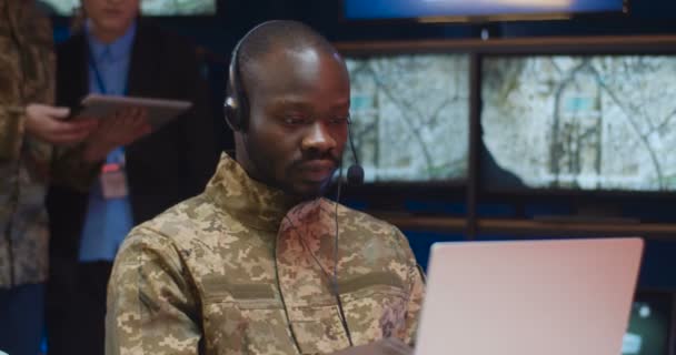 Retrato de homem militar afro-americano em fone de ouvido sentado no computador portátil no centro de controle do exército e sorrindo alegremente para a câmera. Soldado no escritório militar. Oficial na sala de despacho . — Vídeo de Stock