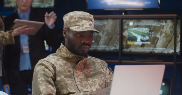 Portret van Afro-Amerikaanse militair in camouflage kleding en pet werkend op latop computer in de controlekamer en glimlachend blij voor de camera. Mannelijke operator bij technisch monitoringcentrum. — Stockvideo