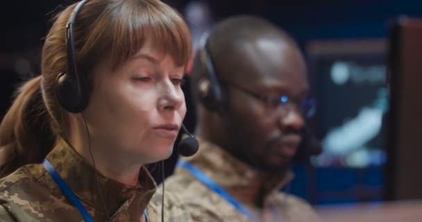 Blanke militaire vrouw in headset zittend achter de computer en pratend via videochat in het controlecentrum van het leger. Vrouwelijke officier spreekt via webcam in het militaire kantoor. Mengvoeders voor gemengde rassen. — Stockvideo